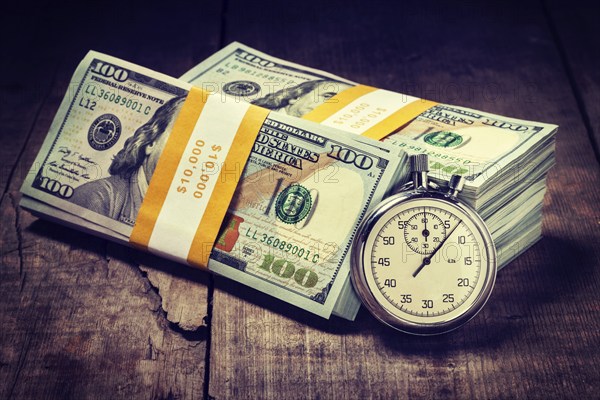 Time is money loan concept background. Vintage retro effect filtered hipster style image of stopwatch and stack of new 100 US dollars 2013 edition banknotes bills bundles on wooden background