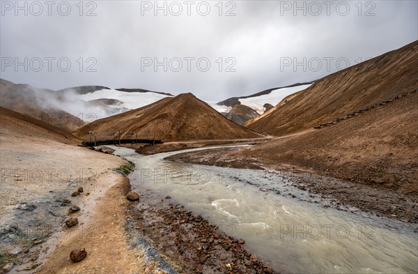 Bridge over steaming stream between colourful rhyolite mountains with snowfields, Hveradalir geothermal area, Kerlingarfjöll, Icelandic highlands, Iceland, Europe