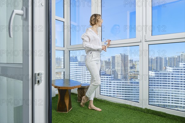 Barefoot woman stands on the balcony of her new apartment and admires the view of the city from the window