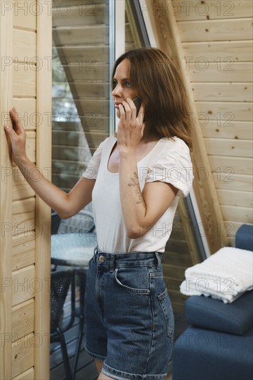 Serious woman inside log a-frame talking on the phone standing by the window