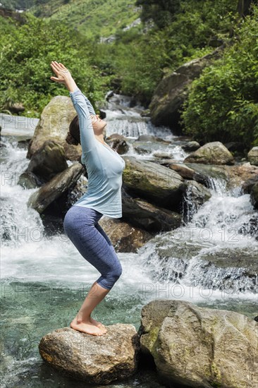 Young sporty fit woman doing yoga asana Utkatasana (chair pose) outdoors at tropical waterfall standing on stone