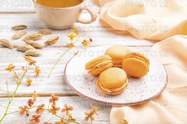 Orange macarons or macaroons cakes with cup of apricot juice on a white wooden background and orange linen textile. Side view, close up