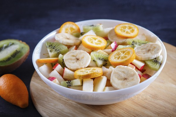 Vegetarian salad of bananas, apples, pears, kumquats and kiwi on black woodem background, close up, selective focus