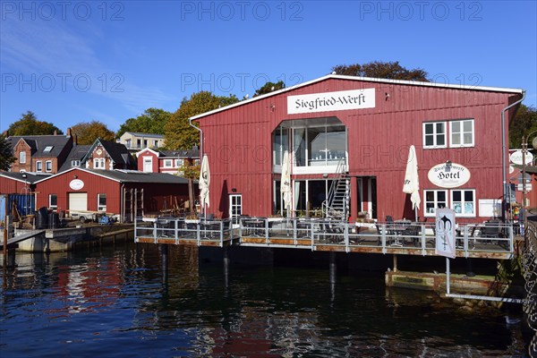 A red wooden building with 'Hotel' lettering on the waterfront under a clear blue sky on a sunny day, Siegfried-Werft, Eckernförde, Schleswig-Holstein, Germany, Europe