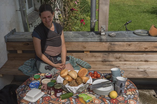 Young mum with baby in a sling at the breakfast table on the terrace, Mecklenburg-Vorpommern, Germany, Europe