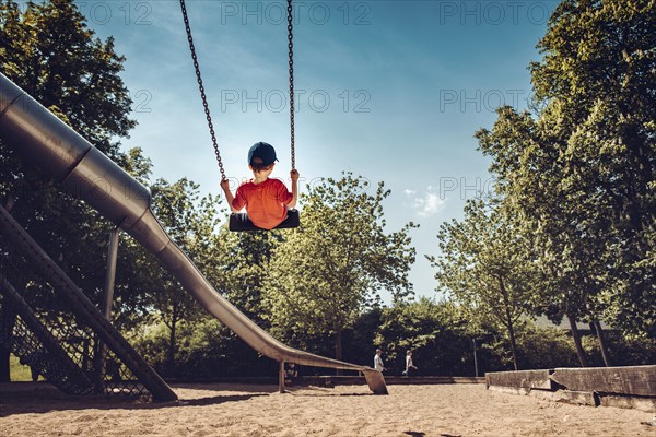 Child swings on playground, surrounded by trees and sunny weather
