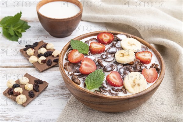 Chocolate cornflakes with milk and strawberry in wooden bowl on white wooden background and linen textile. Side view, close up