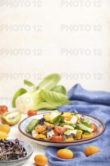 Vegetarian salad of pac choi cabbage, kiwi, tomatoes, kumquat, microgreen sprouts on a white concrete background and blue linen textile. Side view, selective focus, copy space