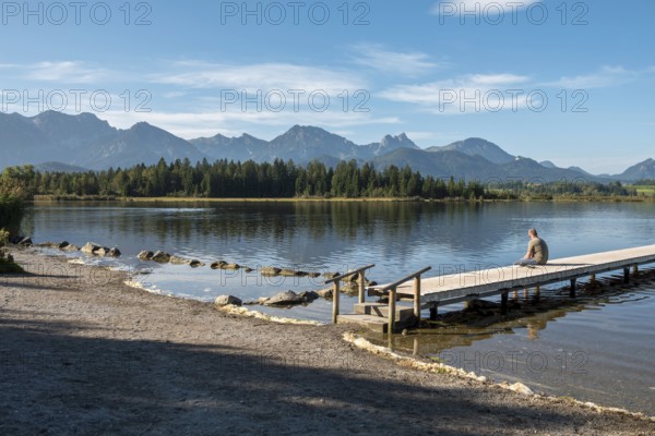 Person sitting on a wooden footbridge looking at a calm lake with mountains in the background, Hopfensee, Hopfen am See, near Füssen, Allgäu Alps, Ostallgäu, Allgäu, Bavaria, Germany, Europe