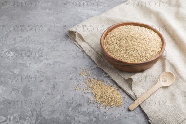 Wooden bowl with raw white quinoa seeds and wooden spoon on a gray concrete background and linen textile. Side view, copy space