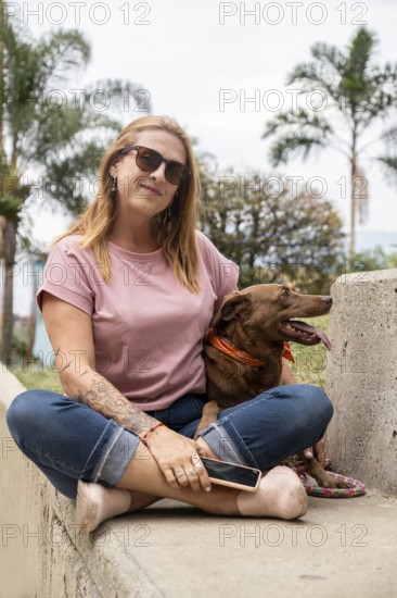 Middle-aged woman with sunglasses, sitting on a cement bench next to her dog, with palm trees in the background