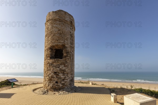 An old tower rises on the beach with sand and sea in the background under a clear sky, Puerco
