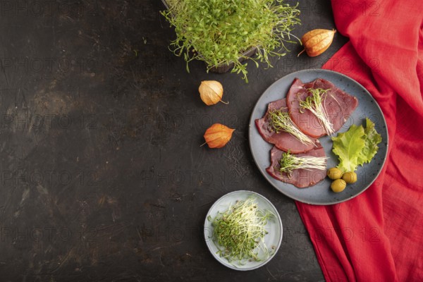 Slices of smoked salted meat with cilantro microgreen on black concrete background and red textile. Top view, flat lay, copy space