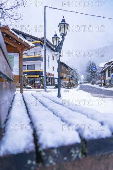 Snow-covered park bench in the centre of a wintry village with traditional houses and street lamps, Enzklösterle, Calw district, Black Forest, Germany, Europe