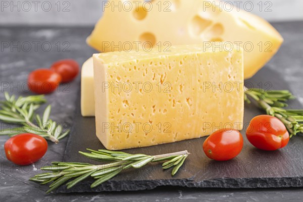 Various types of cheese with rosemary and tomatoes on black slate board on a black concrete background. Side view, close up, selective focus