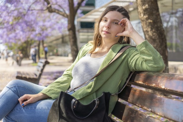 A young woman in a green jacket looks at the camera while sitting relaxed on a bench under purple blooming trees on a sunny day. Blurred people walk in the background, enjoying the pleasant weather