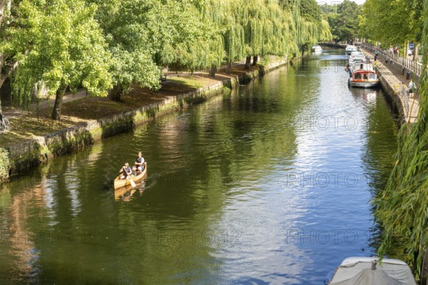 Boats moored people kayaking, River Wensum, Norwich, Norfolk, England, UK treelined riverbank