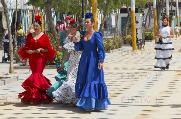 Five woman in different colourful flamenco dresses, with flowers in their hair, strolling on a festive street, Feria de la Manzanilla, Feria de Sanlúcar, Sanlúcar de Barrameda, Sanlucar, Cádiz, Cadiz, Andalusia, Spain, Europe
