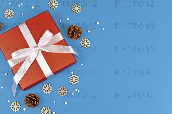 Red Christmas gift box with ribbon surrounded by seasonal pine cones and wooden snowflake ornaments on side of blue background with empty copy space