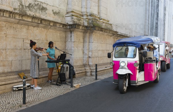 A violinist and a singer play music in front of an old wall while a pink tuk-tuk drives by, Old Town, Lisbon, Lisboa, Portugal, Europe