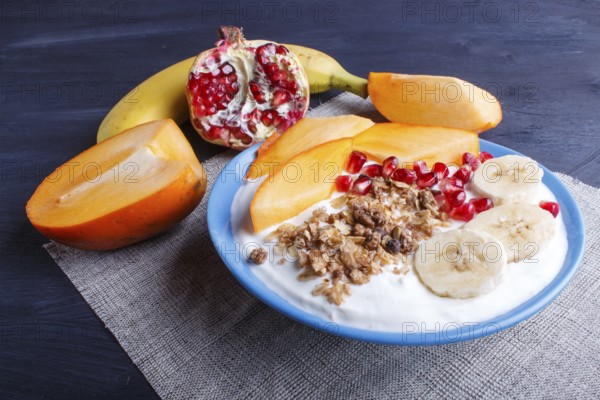 A blue plate with greek yogurt, granola, persimmon, banana, pomegranate black wooden background. close up