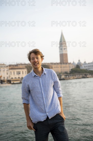 Young man in striped shirt and shorts on the banks of the Grand Canal, looking into the camera, Campanile in the background, Venice, Veneto, Italy, Europe