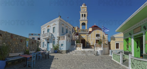 Sunny Plati Square in the picturesque village of Olymbos with colourful buildings and the church of Kimisi tis Panagías, decorated with Greek flags, Olymbos, Karpathos, Greece, Europe