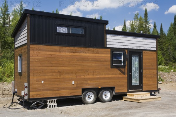 Contemporary brown stained pine and grey oak wood shingles clad cottage style 8 x 24 foot sloped roof mobile Mini House facade with black trimmed windows and entrance door in summer, Quebec, Canada, North America