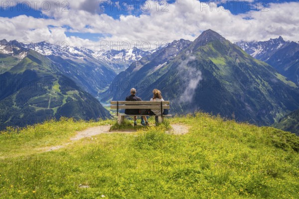Two people sit on a bench and look out over a valley with majestic mountains in the background, Penken, Zillertal. Austria