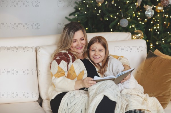 Happy family grandmother and little girl daughter sitting together on sofa and reading book while spending time together at home during Christmas holidays in winter, hugging and covering with wool plaid