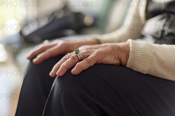An elderly woman sits on a couch with her hands resting on her knees, wearing a ring on her left hand. Loneliness and old edge concept