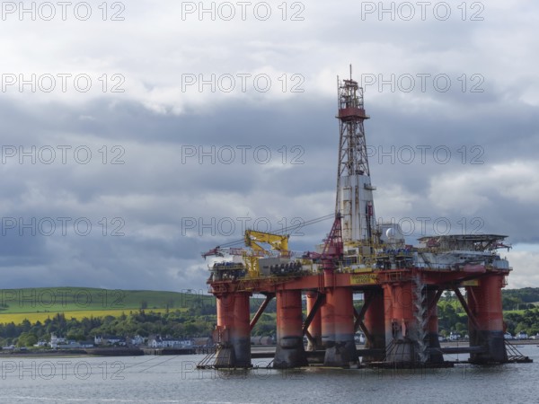 An oil drilling platform stands in the calm sea, behind it a cloudy sky and green landscape, drilling platform in red against a blue sky in the north sea, inverness, Scotland, Great Britain