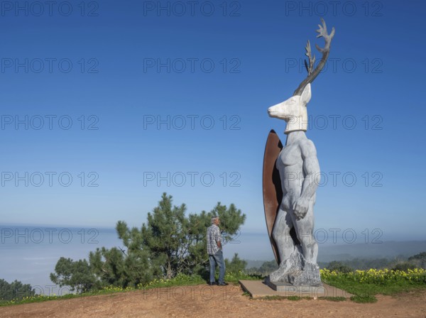 A man stands in front of a surfer statue with a deer head, Nazaré, Portugal, Europe