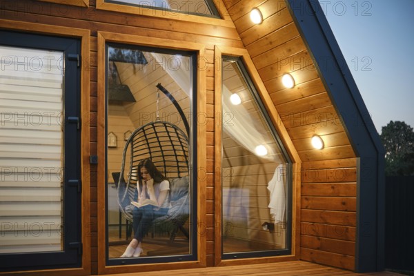 Woman relaxing in a hanging chair with a book inside a stylish wooden cabin. The warm lighting and large windows create an inviting atmosphere, perfect for relaxation