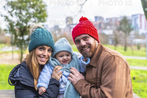 Smiling parents holding their baby, wearing winter clothes and wool hats, having fun together in a city park
