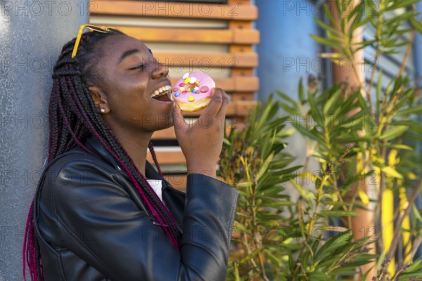 Cheerful African woman with leather jacket biting a doughnut in the corner of street