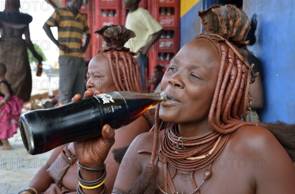 Traditionally dressed Himba woman drinking a coke in front of a bar, Opuwo, Kaokoland, Kunene, Namibia, Africa