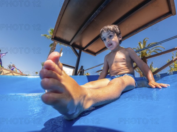Young boy enjoying a water slide at a water park during summer vacation, surrounded by palm trees and a clear mediterranean sky in alicante