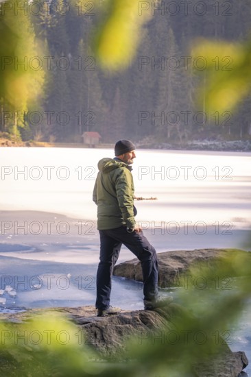 Person standing on the shore of a frozen lake, framed by rocky shore and forest, Glaswaldsee, Bad Rippoldsau-Schapbach, district of Wolfach, Black Forest, Germany, Europe