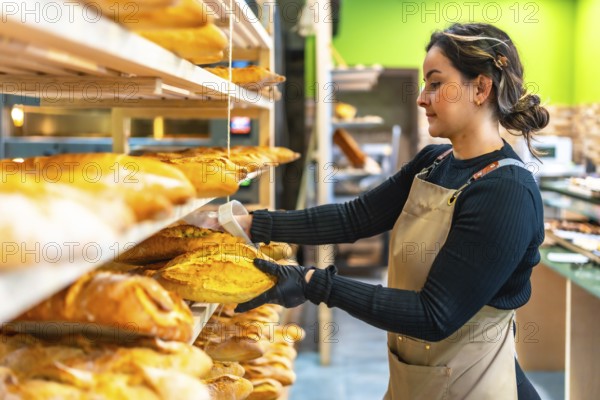 Side view of a young latin woman worker arranging artisan bread from shelf in the bakery shop