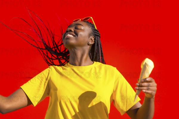 Cool happy african woman dancing and eating ice cream under the sun and against a red wall