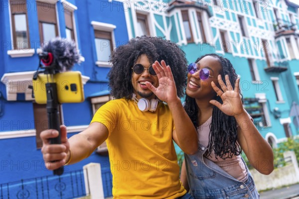 African couple broadcasting a life video waving at camera next to blue colorful house