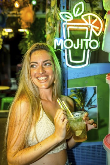 Vertical photo of a happy woman smiling while drinking mojito cocktail in a bar enjoying summer night