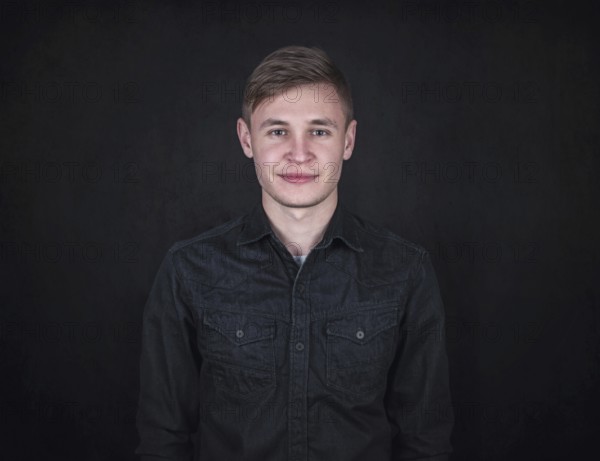 Portrait of a carefree young man against a black wall background looking to camera. Relaxed male model with joyful face expression wearing dark blue jeans jacket
