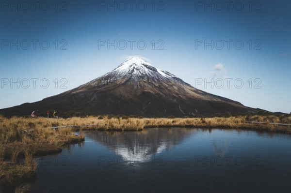 A snow-capped mountain is reflected in a still lake while people walk along the shore, Mount Taranaki, New Zealand, Oceania