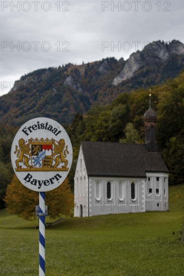 Free State of Bavaria sign with Nussdorf-Windshausen Kreuzkirche in the background, autumnal mountain landscape (Heuberg) with overcast sky, surrounded by trees and a green meadow, Nussdorf, Bavaria, Germany, Europe