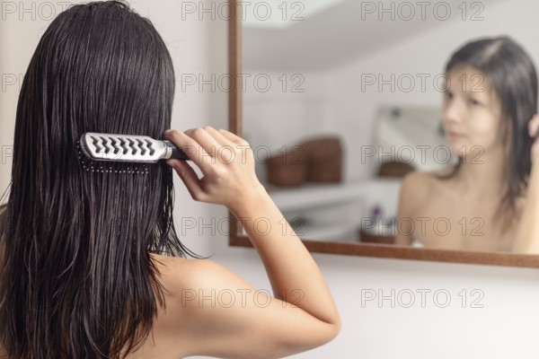 Young woman standing looking at her reflection in the bathroom mirror while brushing her wet hair with a hairbrush after washing it, view from behind