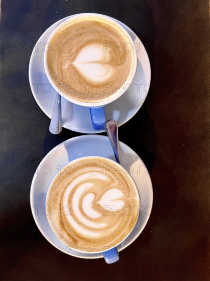 Top-down view of two cappuccinos with heart-shaped latte art, served in blue cups on a dark table.