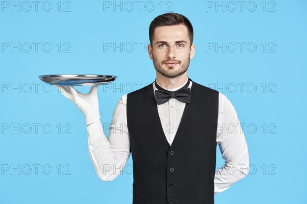Attractive young waiter holding empty silver tray over blue background with copy space. Restaurant, horeca, first class service concept