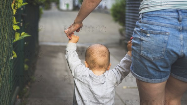 Learning to walk concept: Toddler is holding the hand of his mother, making his first steps. Close up from behind